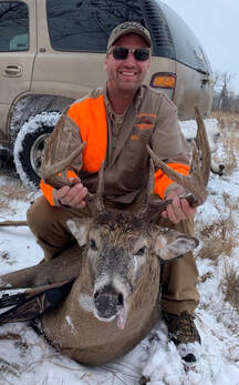 Chad in orange vest with a harvested whitetail buck in a snowy field